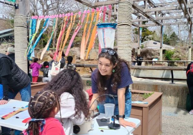 Woman talking to children looking through microscope, colored banners behind her.